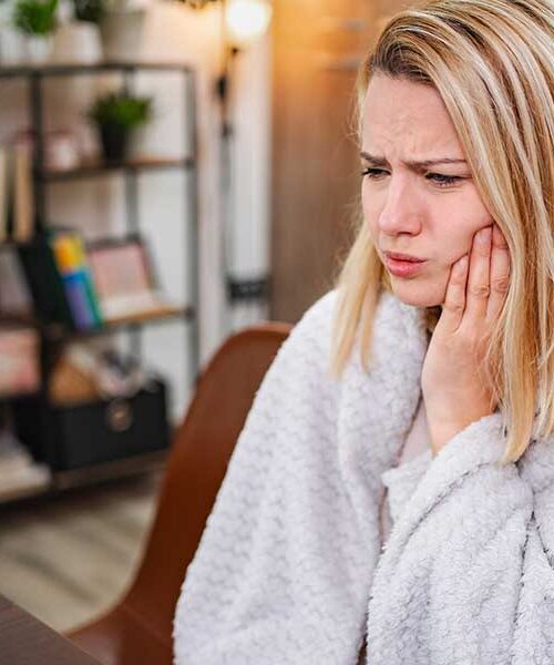 A woman in a robe sitting at a table with coffee, holding her cheek in discomfort due to morning jaw pain.