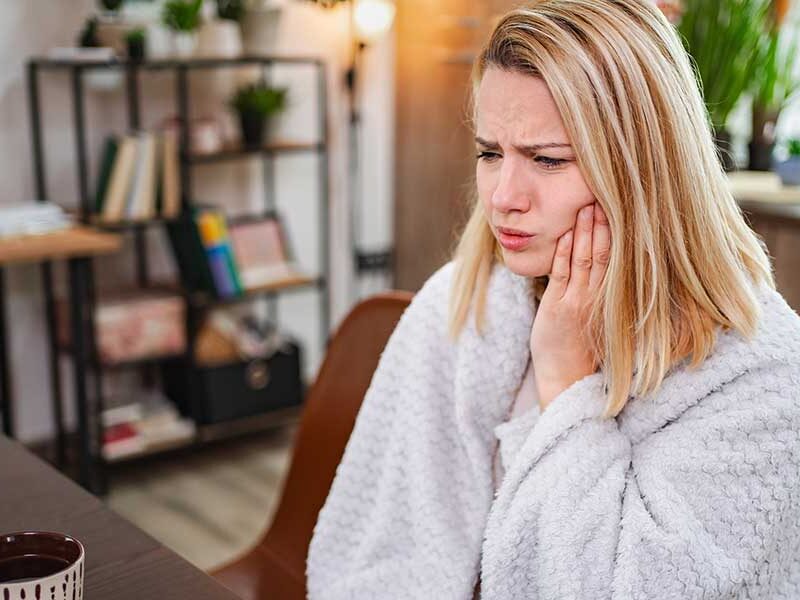 A woman in a robe sitting at a table with coffee, holding her cheek in discomfort due to morning jaw pain.