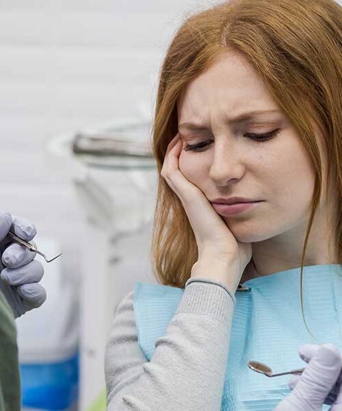 A woman holding her aching cheek in a dental chair while a dentist holding dental instruments evaluates her severe toothache.
