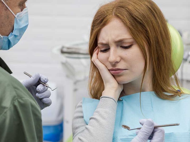 A woman holding her aching cheek in a dental chair while a dentist holding dental instruments evaluates her severe toothache.